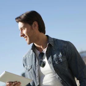 Stylish young man in denim jacket outdoors, holding a tablet. Sunny day.