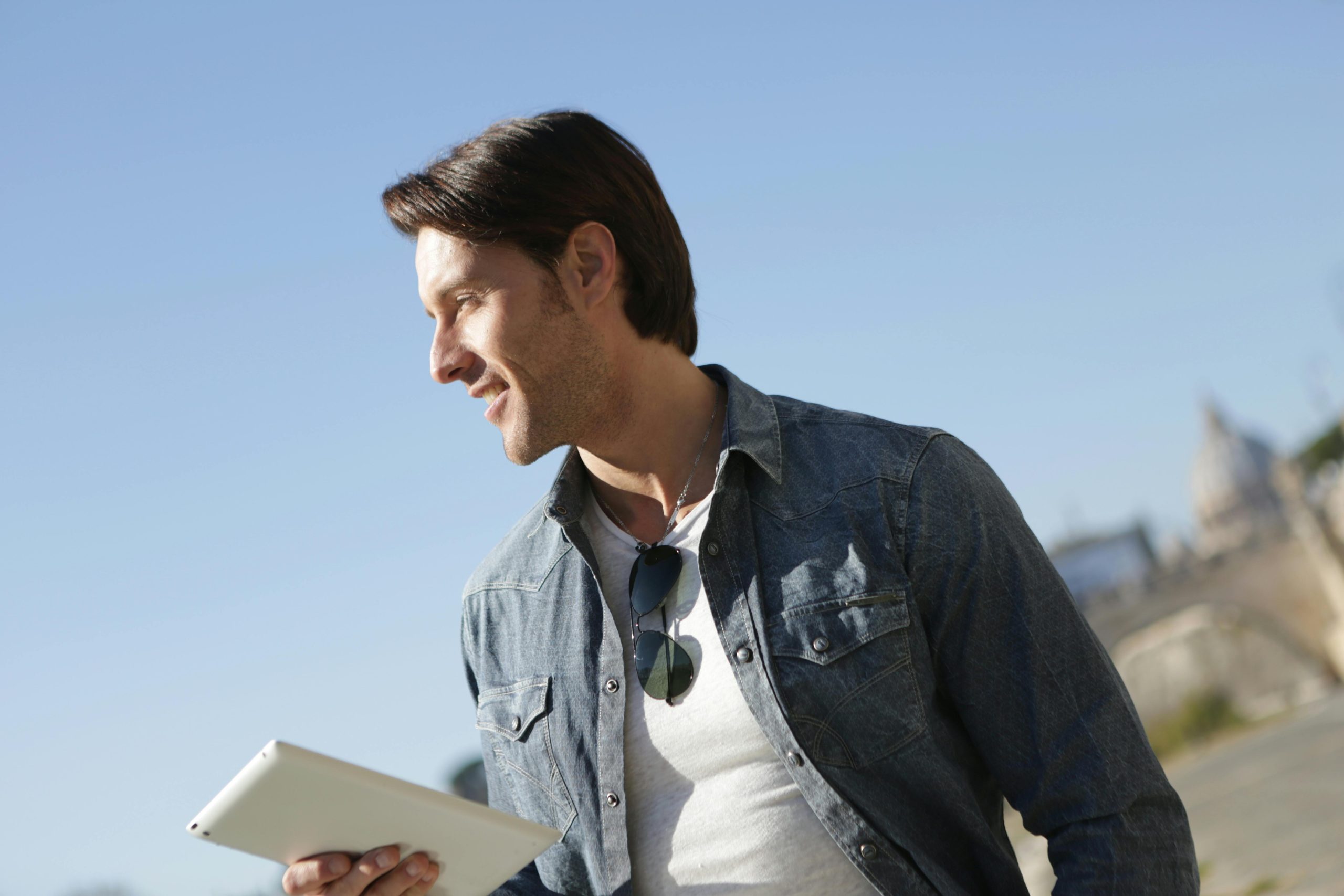 Stylish young man in denim jacket outdoors, holding a tablet. Sunny day.